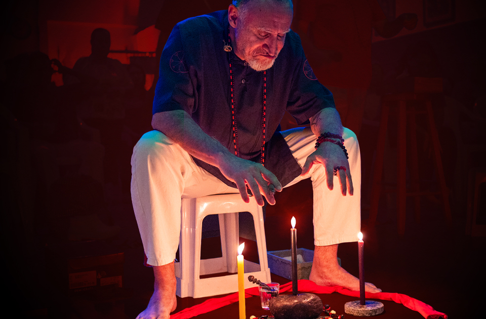 Homem idoso, de barba branca, sentado em um banquinho em um ambiente escuro e iluminado por velas. Ele veste uma camisa escura com símbolos esotéricos e um colar de contas vermelhas. Diante dele, sobre um pano vermelho no chão, há velas acesas, pedras e outros elementos ritualísticos. Sua expressão séria e sua postura meditativa sugerem concentração em um ritual espiritual.