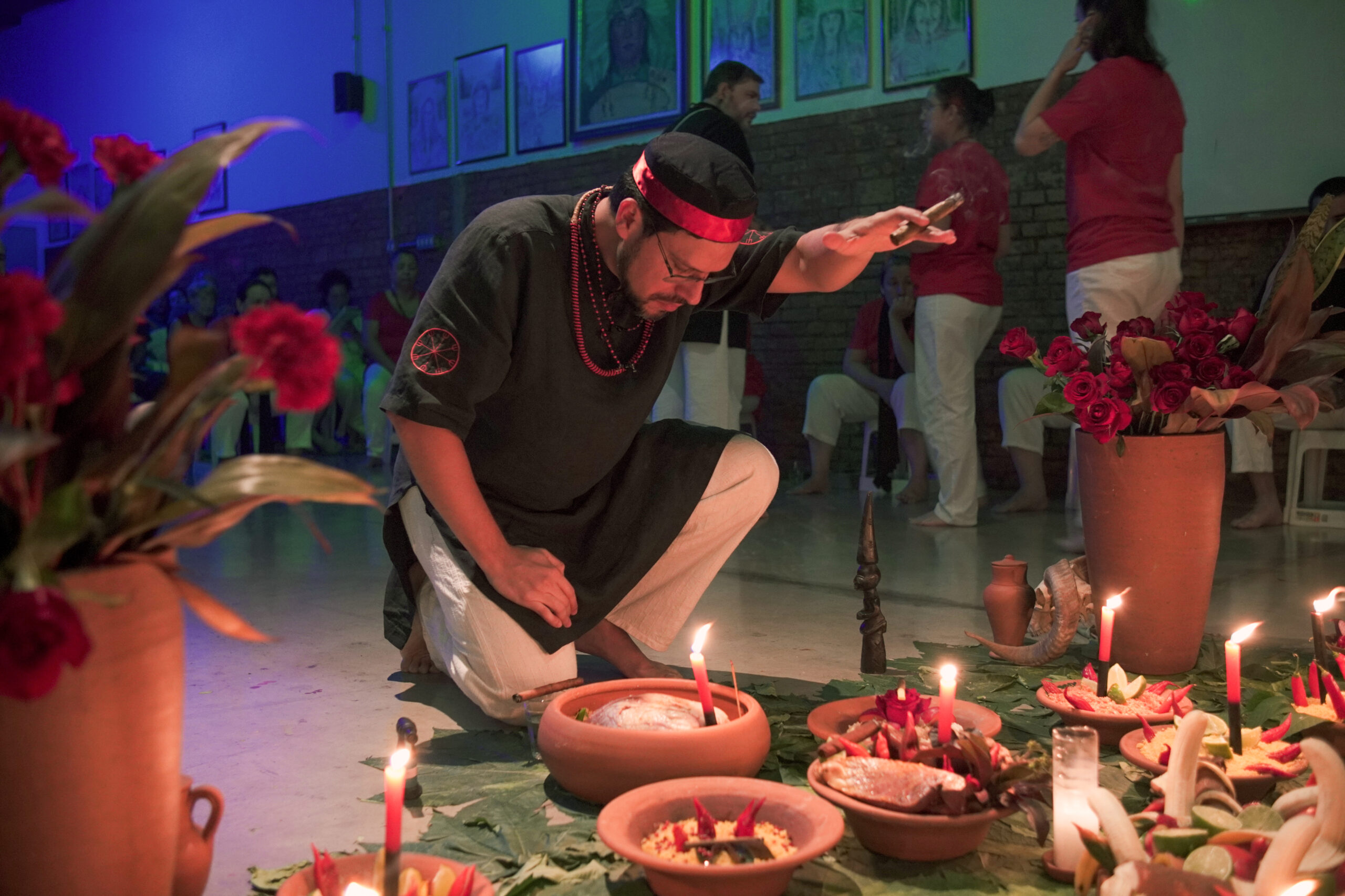 Homem ajoelhado em um ritual de Umbanda, vestindo roupa preta com detalhes vermelhos e segurando um charuto. À sua frente, oferendas em recipientes de barro, decoradas com velas, folhas, pimentas e outros elementos sagrados. Ao fundo, participantes vestindo roupas brancas e vermelhas observam o ritual em um ambiente iluminado por velas e decorado com quadros na parede de tijolos.
