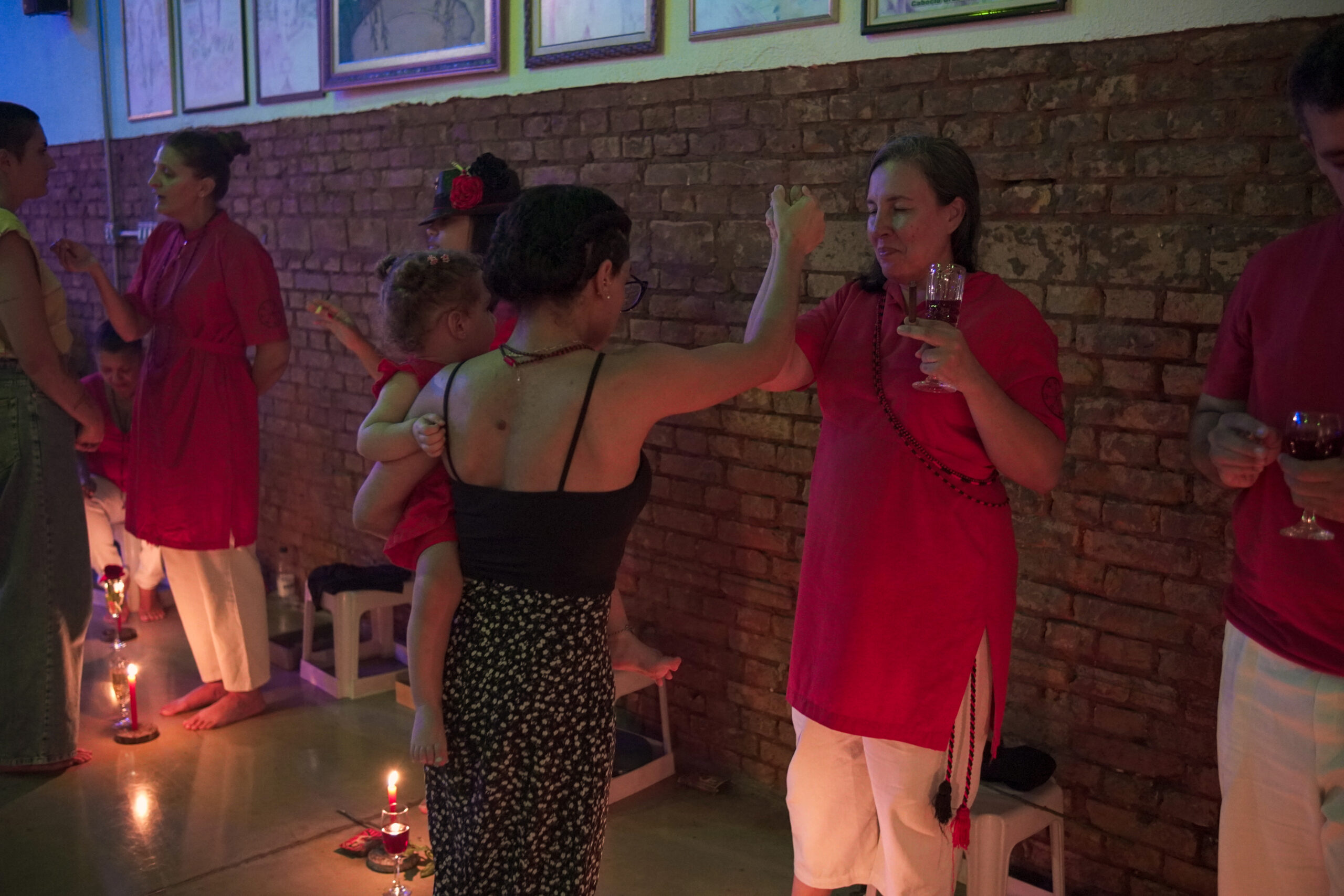 Gira de Pombagira com a presença de mulheres vestidas de vermelho e branco, em um ambiente iluminado por velas e decorado com quadros na parede de tijolos. Uma mulher segura uma taça de vinho enquanto recebe um gesto ritualístico de outra mulher que carrega uma criança no colo. O clima da cena é espiritual e comunitário.